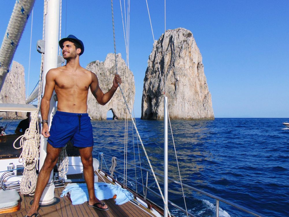 Young beautiful man standing in swimming suit on a yacht near blue sea water of Capri